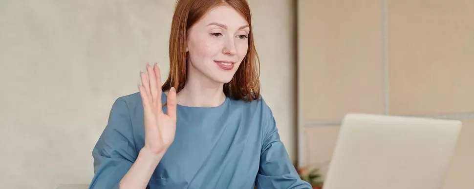 Woman in blue shirt waving at laptop during video call.