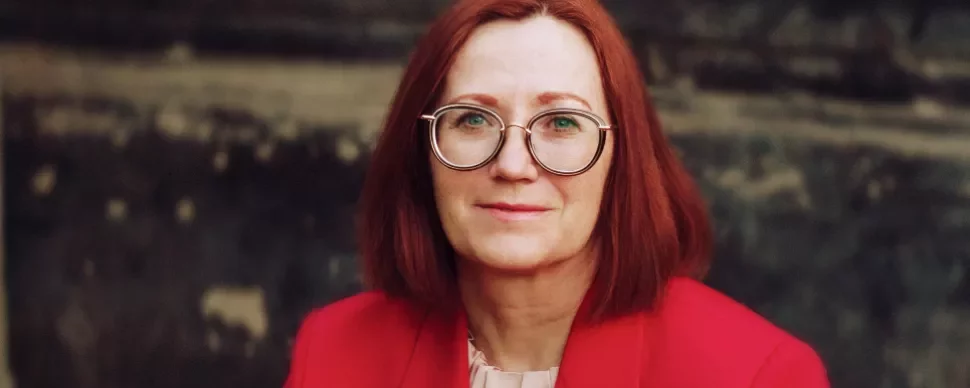 Woman with red hair and glasses wearing a red jacket.