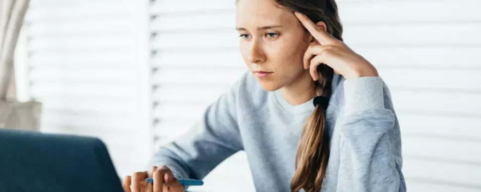 Young woman focuses intently on laptop screen, holding pen.