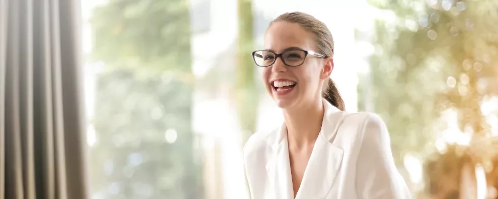 Woman in glasses laughing while sitting in a bright room.