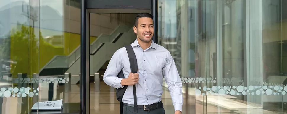 Smiling man with a backpack exits a glass building entrance.