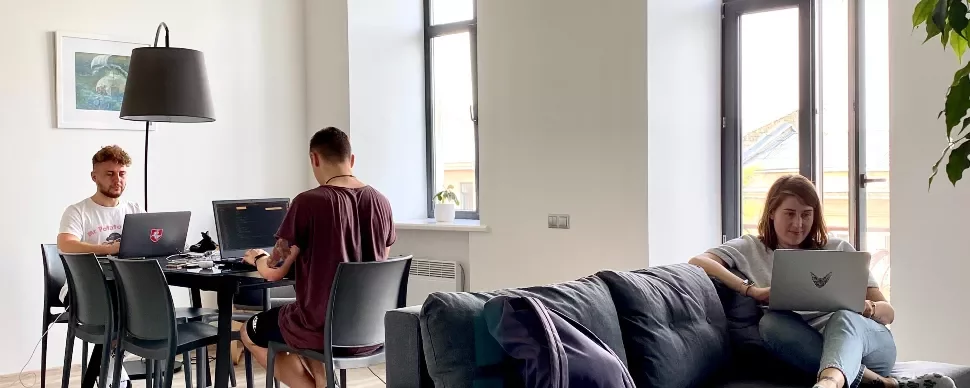 Three people working on laptops in a bright room with large windows.
