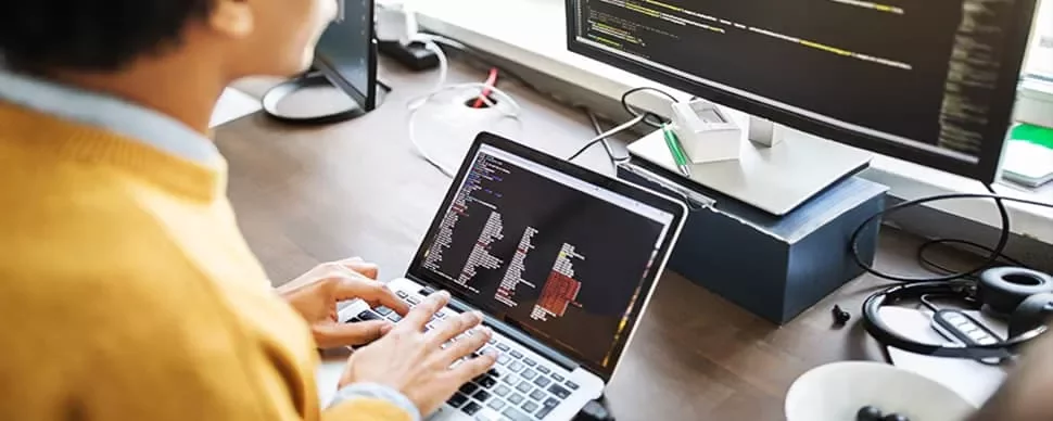 Person coding on a laptop with a larger screen displaying code.