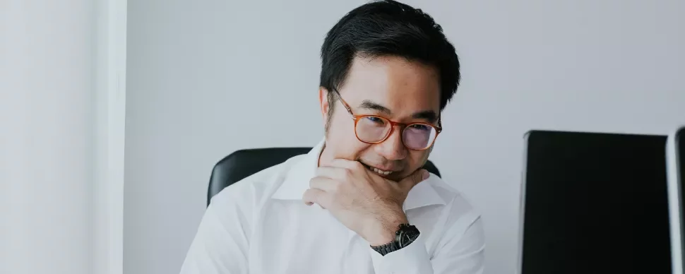 Man in glasses smiling at a desk with two computers.