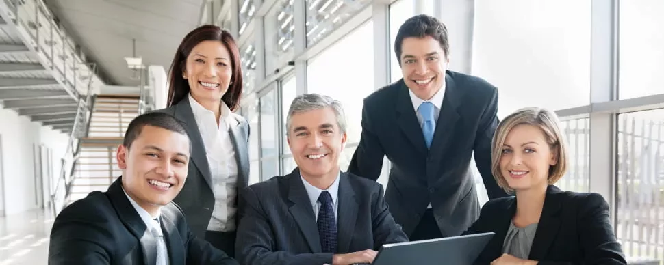 A diverse group of business professionals smiling around a laptop in a modern office.