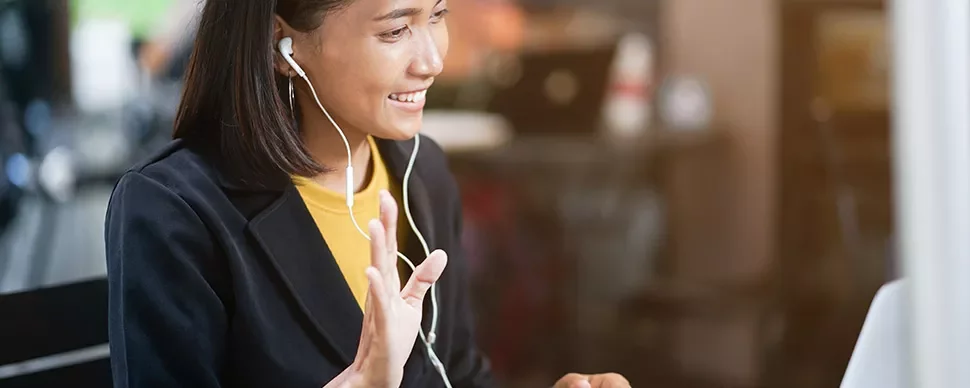 Woman smiling and waving during a video call, wearing earphones, with a blurred background.