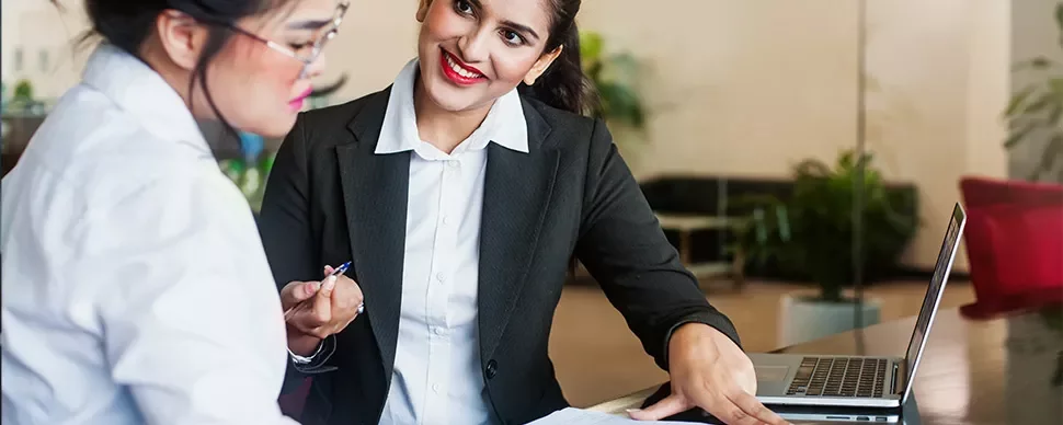 Two women having a discussion at a desk in an office setting.