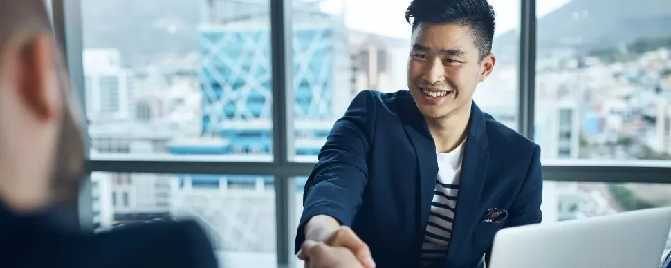 Two people shaking hands in an office with city view in background.