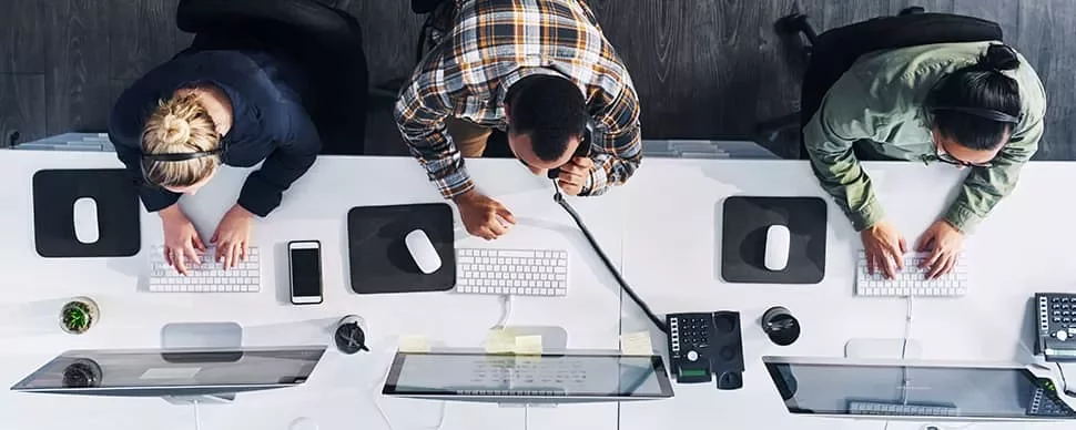 Overhead view of three people working at office desks with computers, using headsets and keyboards.