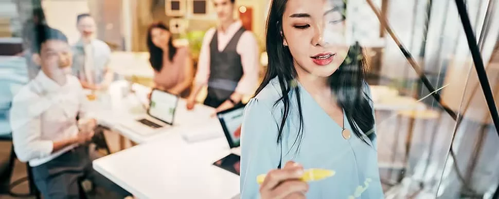 Woman writing with marker on glass, with colleagues in conference room.
