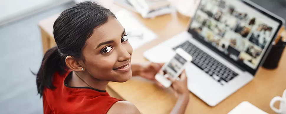 Woman smiling while holding phone at desk with laptop open.