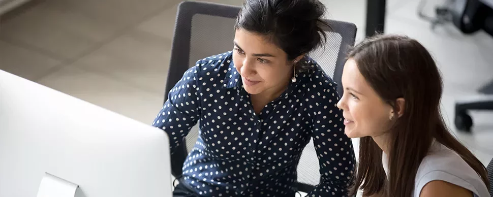 Two women smiling and working together at a computer desk in an office setting.