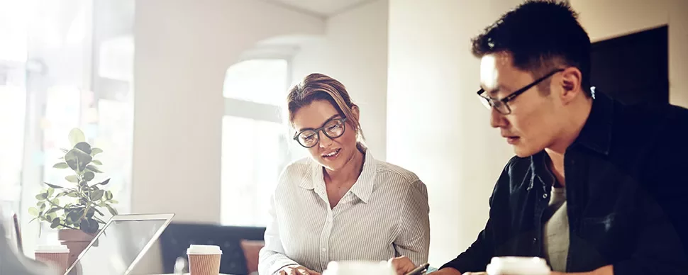Two people working together on documents at a table with coffee and a laptop.