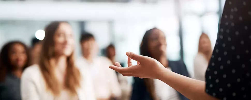 A speaker gesturing in front of a blurred audience.