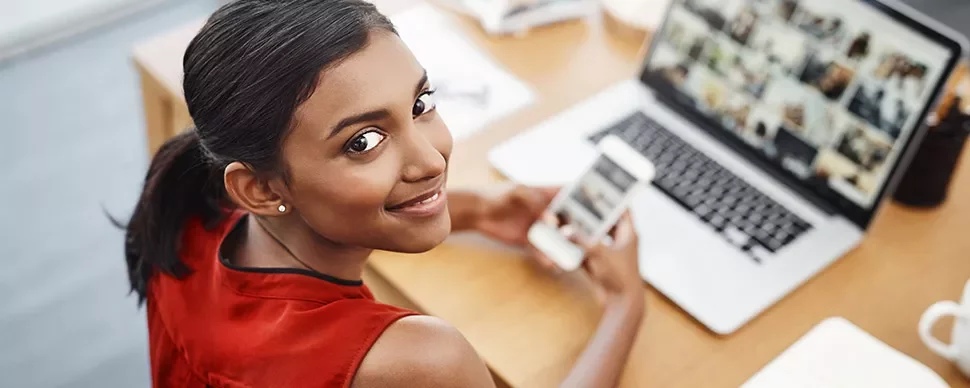 Woman in red top smiling, holding a phone, looking at a laptop with images.