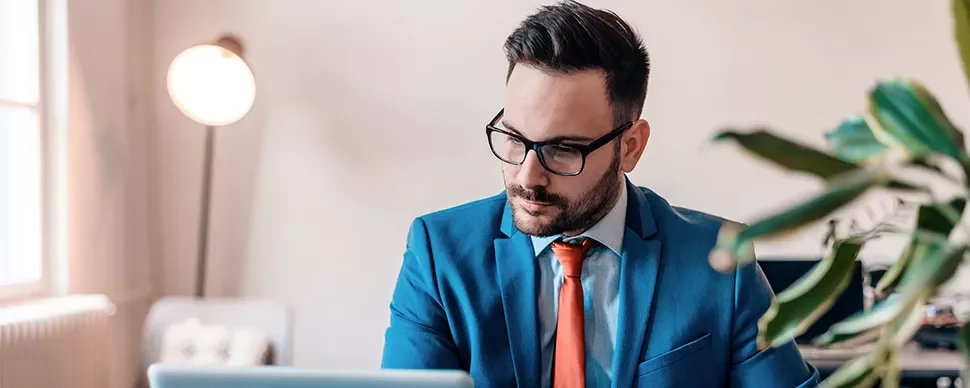 Man in blue suit and glasses working on a laptop in a modern office.