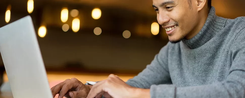 Man in gray sweater smiling while using a laptop.