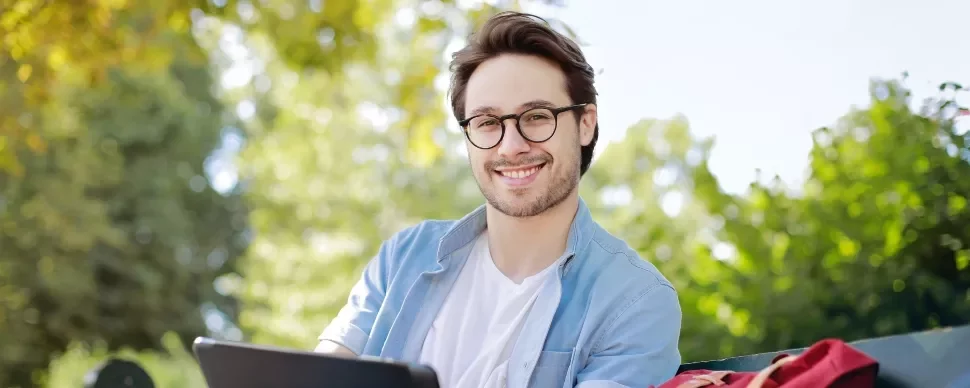 Smiling person with glasses sitting outdoors, holding a tablet.