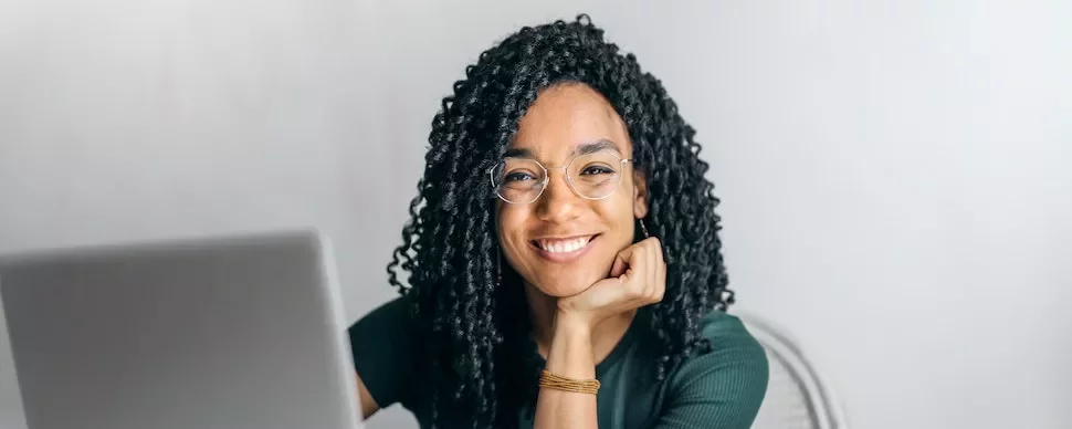 Woman with glasses smiling at laptop, hand on chin.