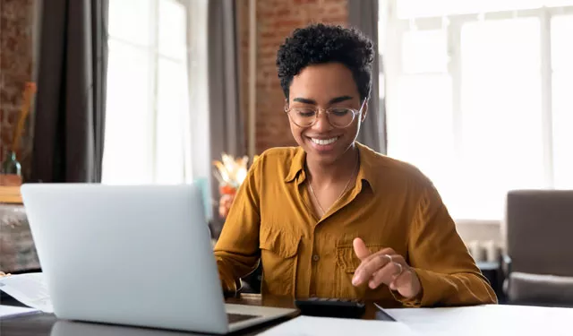 Person smiling and using a calculator while working on a laptop at a desk.