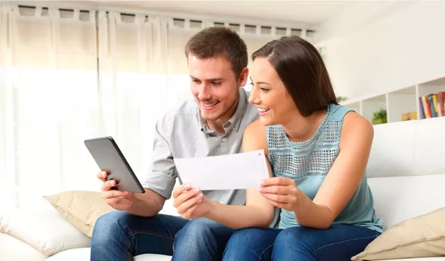 Couple sitting on a couch, smiling at a tablet and holding a piece of paper.