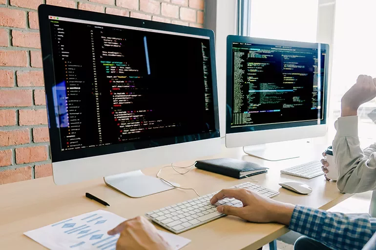 Two computer monitors displaying code in an office setting with two people working at a desk.