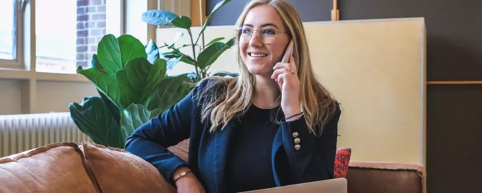 Smiling woman in glasses talking on the phone, sitting on a couch with a laptop nearby.