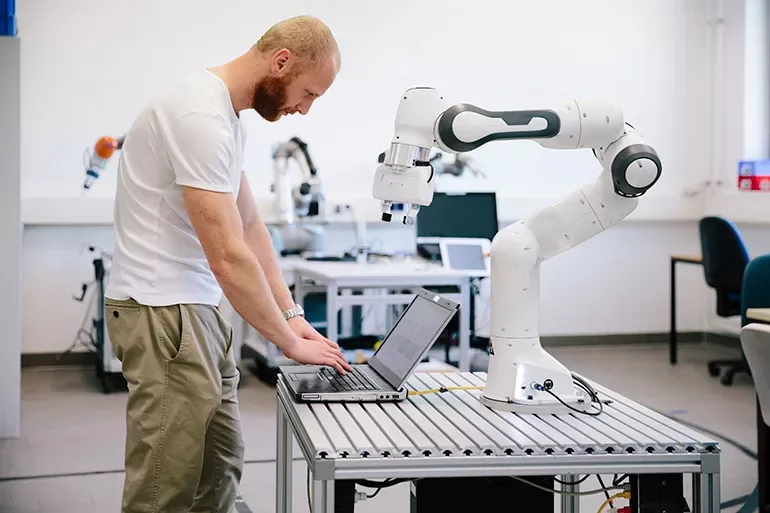 Engineer working on a laptop next to a robotic arm in a lab.
