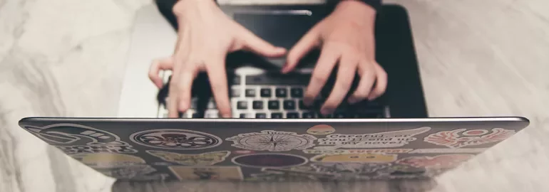 Hands quickly typing on a laptop covered with various colorful stickers.
