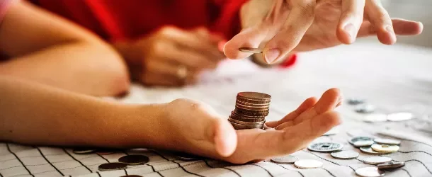 Hand holding a stack of coins, with another hand adding a coin to the stack on a table.