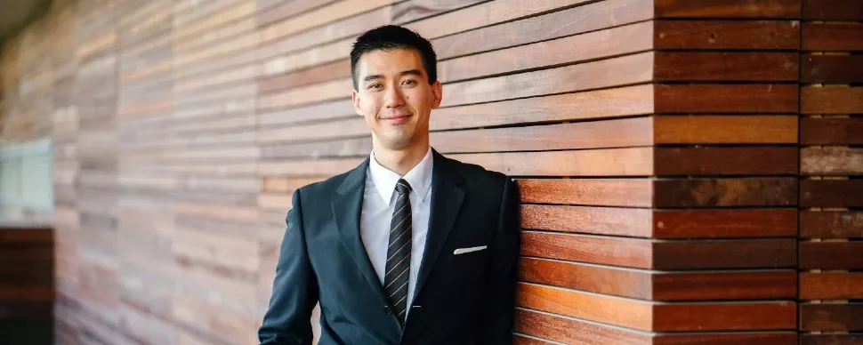Man in a suit standing by a wooden wall, smiling.