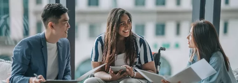 Three people in a meeting, smiling and discussing with papers and tablets.