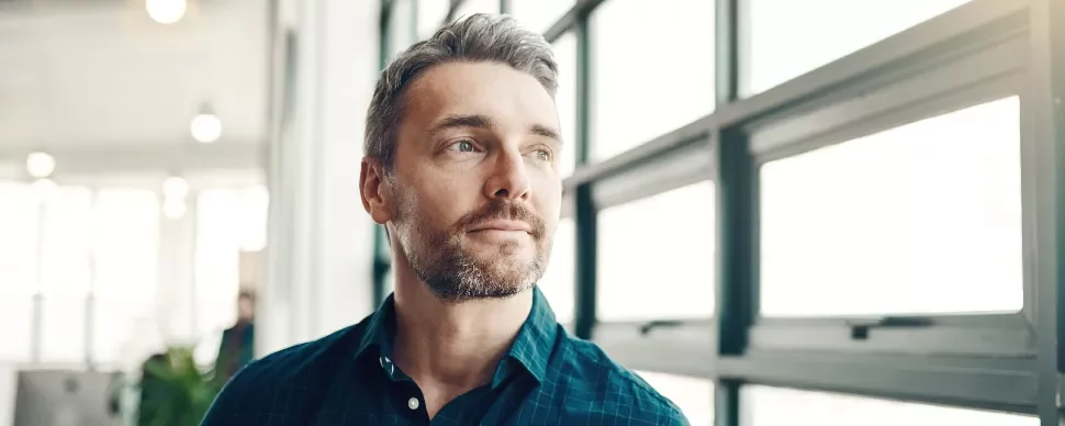 Man with a beard looking out a large window, wearing a blue shirt.