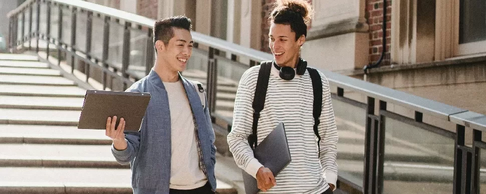 Two students with laptops walking and talking outside.