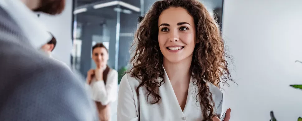 Person with curly hair smiling in office setting, two colleagues in the background.