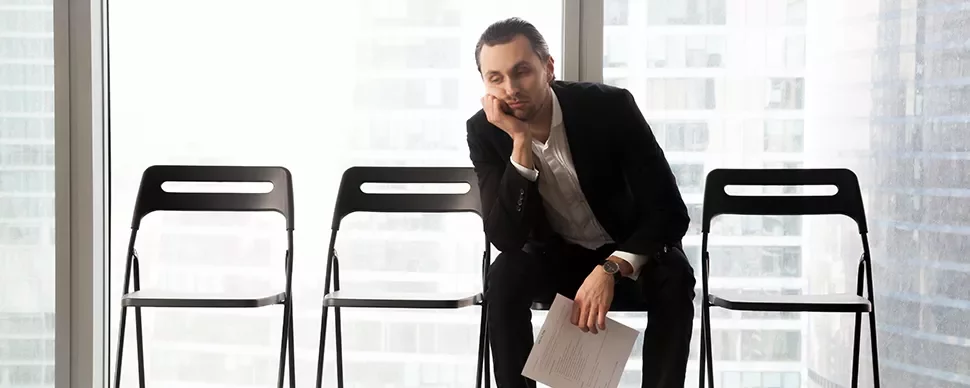 Man in a suit sitting on a chair, resting his head on his hand, looking tired, with empty chairs beside him.