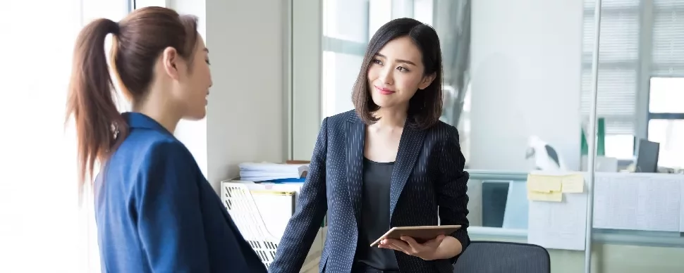 Two women in business attire having a conversation in an office.