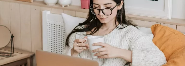Woman with glasses sipping coffee while using a laptop.