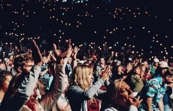 A large crowd cheering at a concert, with lights in the background.
