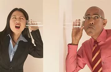 Two people using cups to listen through a wall.