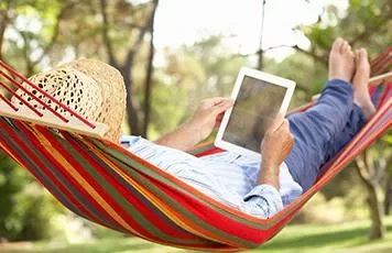 Person relaxing in a colorful hammock with a tablet on a sunny day.