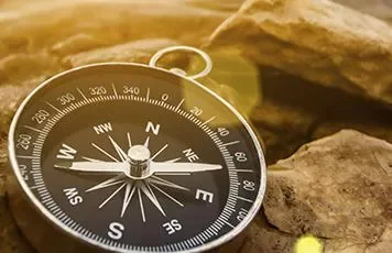 A compass resting on rocky ground, showing cardinal directions with sunlight casting a warm glow.