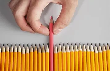 A hand picking a pink pencil out of a row of yellow pencils.