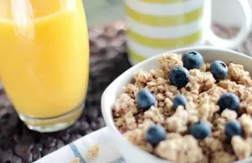 Bowl of granola with blueberries next to a glass of orange juice and a striped mug.
