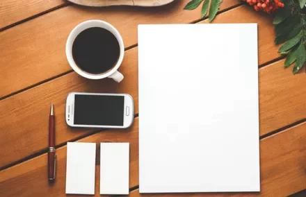 Workspace with coffee, smartphone, pen, and blank paper on a wooden table.