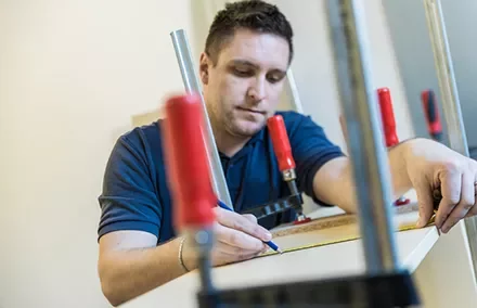 Man measuring and marking a wooden board using a pencil and clamps.