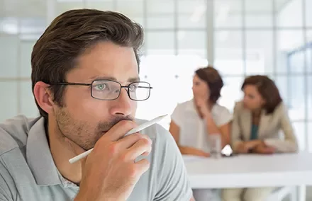 Man thinking with pen in hand, two women in background.