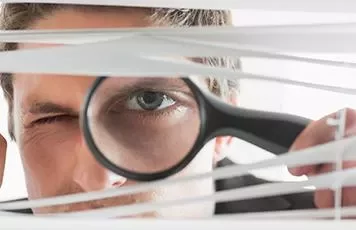 Person looking through blinds with a magnifying glass.