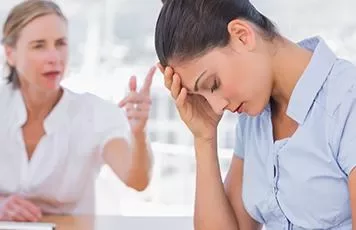 A woman sits with her head in her hand while another woman speaks to her in the background.