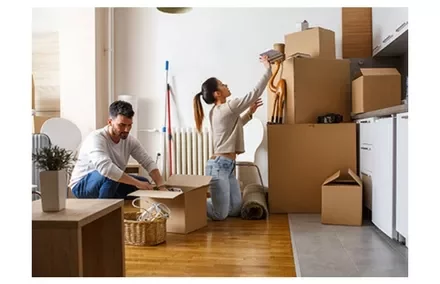 A couple unpacking boxes in a room with wooden floors.